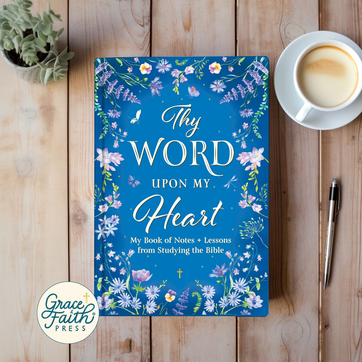 Blue book titled 'Thy Word Upon My Heart' with floral design on a wooden surface with a cup of coffee and pen.